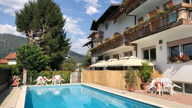A pool area with lounge chairs and umbrellas, surrounded by a building with balconies and potted plants. - Albergo Gilda (Caldonazzo)