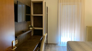 A hotel room with a bed, a TV mounted on the wall, a small table with a plant, and a window with sheer curtains. - Albergo Gilda (Caldonazzo)