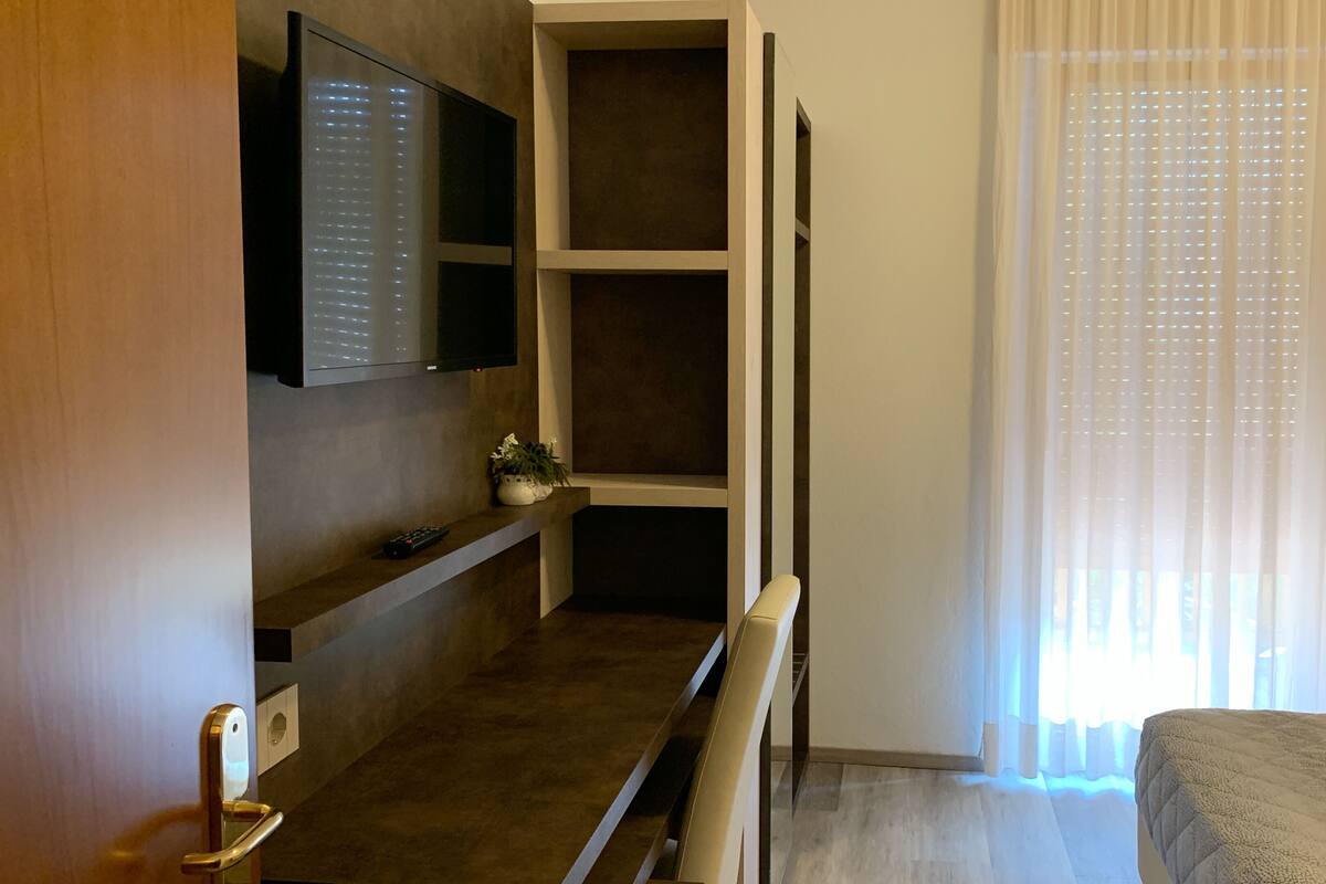 A hotel room with a bed, a TV mounted on the wall, a small table with a plant, and a window with sheer curtains.