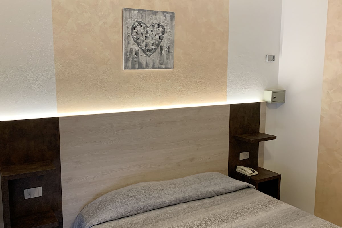 A hotel room with a bed, bedside tables, a wall-mounted light, and a decorative heart-shaped artwork.