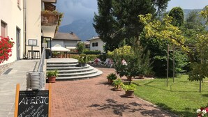 A hotel entrance with a paved pathway leading to a seating area with outdoor chairs and a table. There are potted plants and a signboard visible. - Albergo Gilda (Caldonazzo)