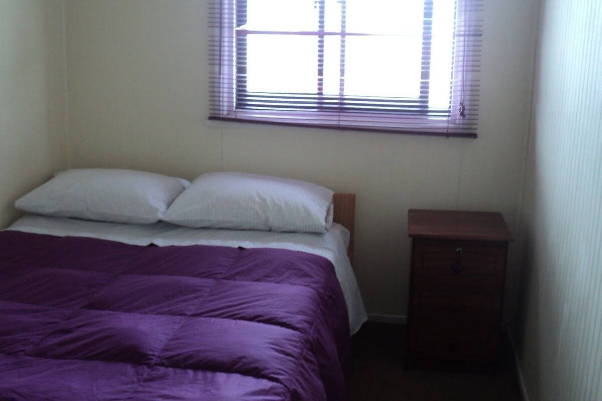 A small, simple bedroom with a bed, a nightstand, and a window with blinds.
