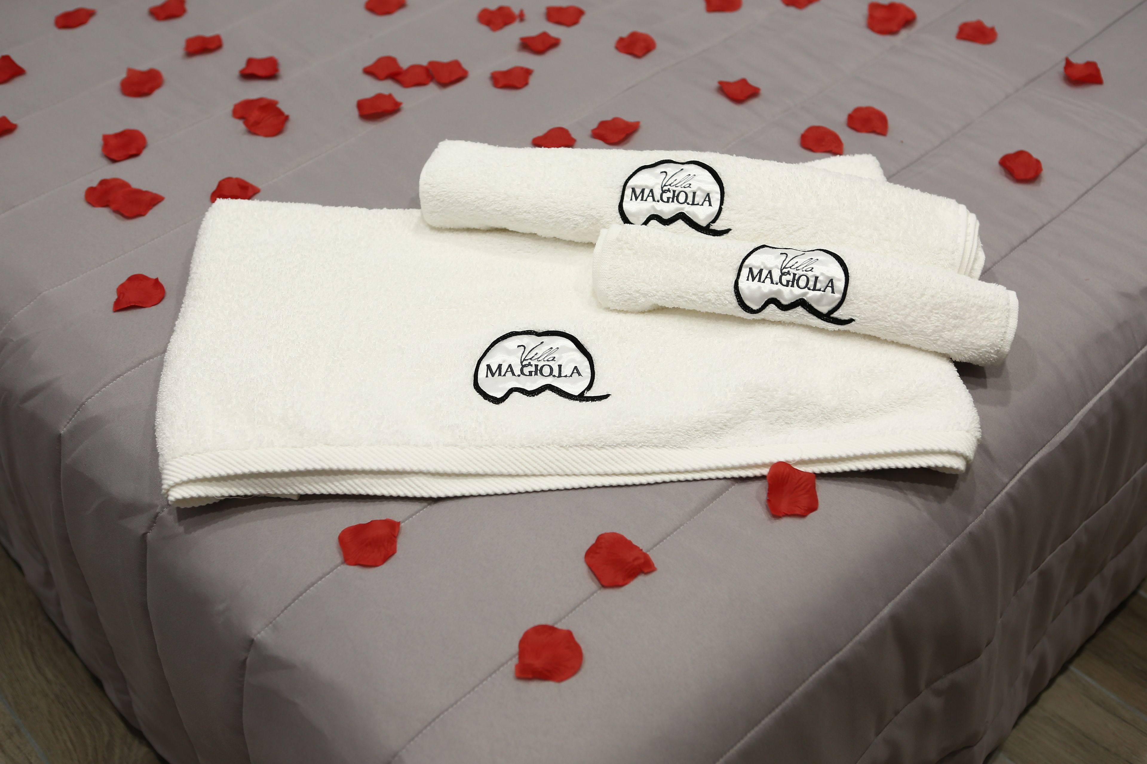 A neatly folded white towel with "Villa Magiola" embroidered on it, placed on a bed with a grey bedspread and scattered red rose petals.