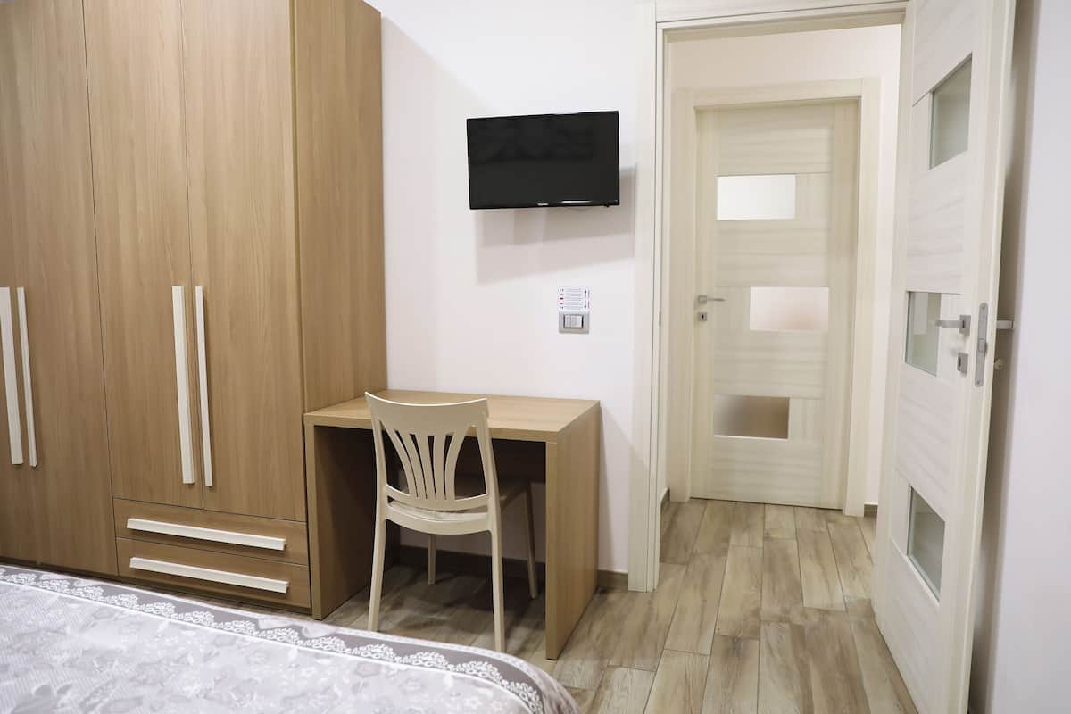 A bedroom with a wooden wardrobe, a desk with a chair, a television mounted on the wall, and a door with frosted glass panels.