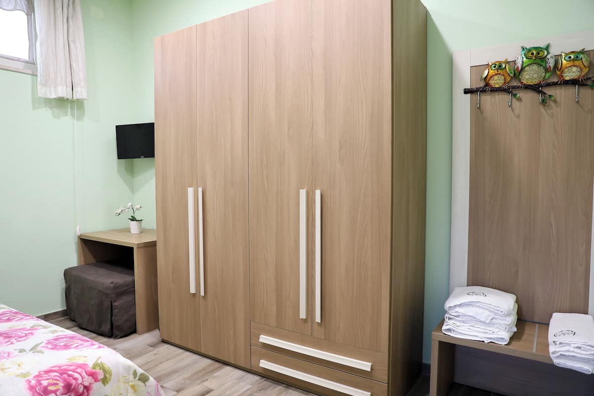 A bedroom with a large wooden wardrobe, a small bedside table with a plant, and a shelf with folded towels.