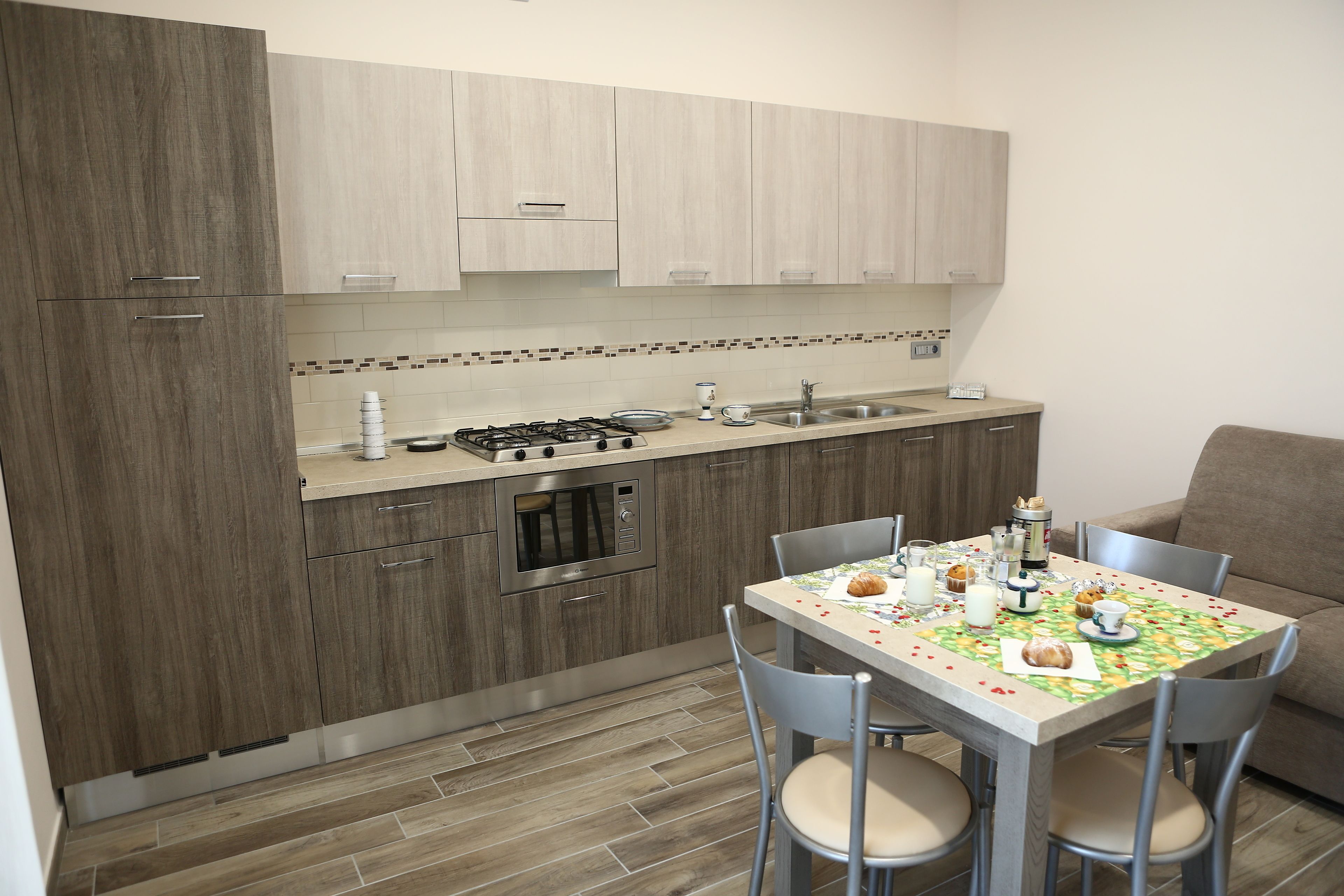 Een kleine keuken met houten kastjes, een gasfornuis, een magnetron en een spoelbak. In de aangrenzende woonkamer staat een eettafel met stoelen en een beige bank.