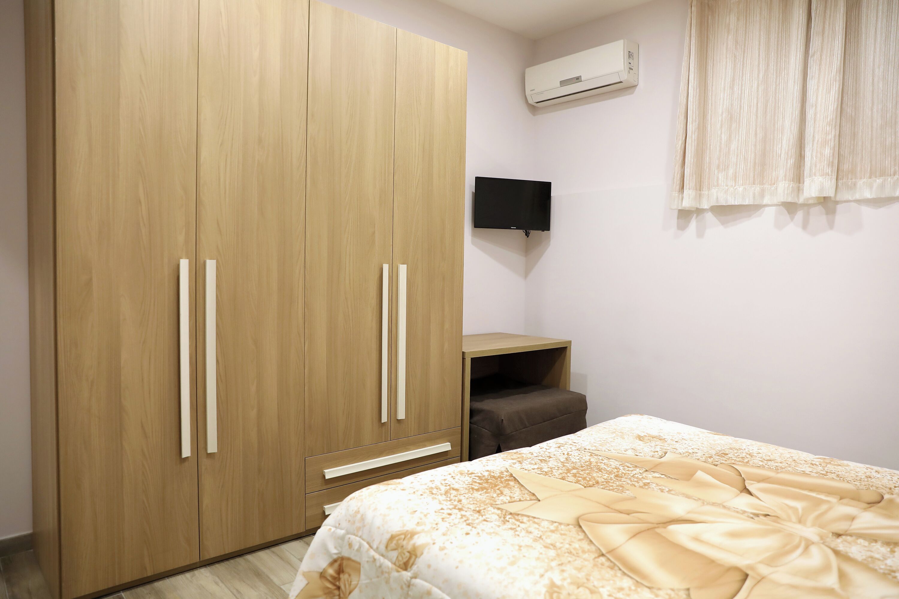 Une chambre avec une armoire en bois, un téléviseur à écran plat fixé au mur, un lit recouvert d’une couvre-lit à motifs et une petite table de chevet.