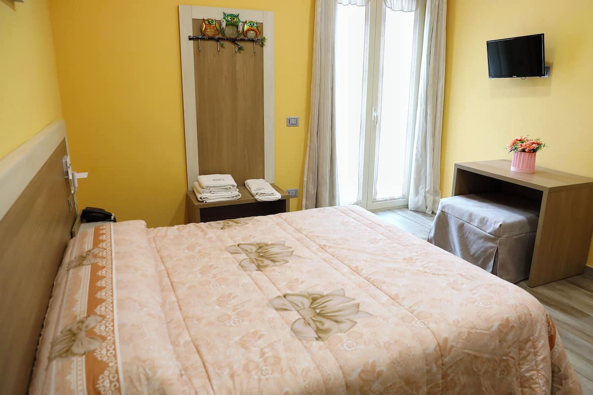 A bedroom with a bed, a wooden wardrobe, a television on the wall, and a small table with a vase of flowers.