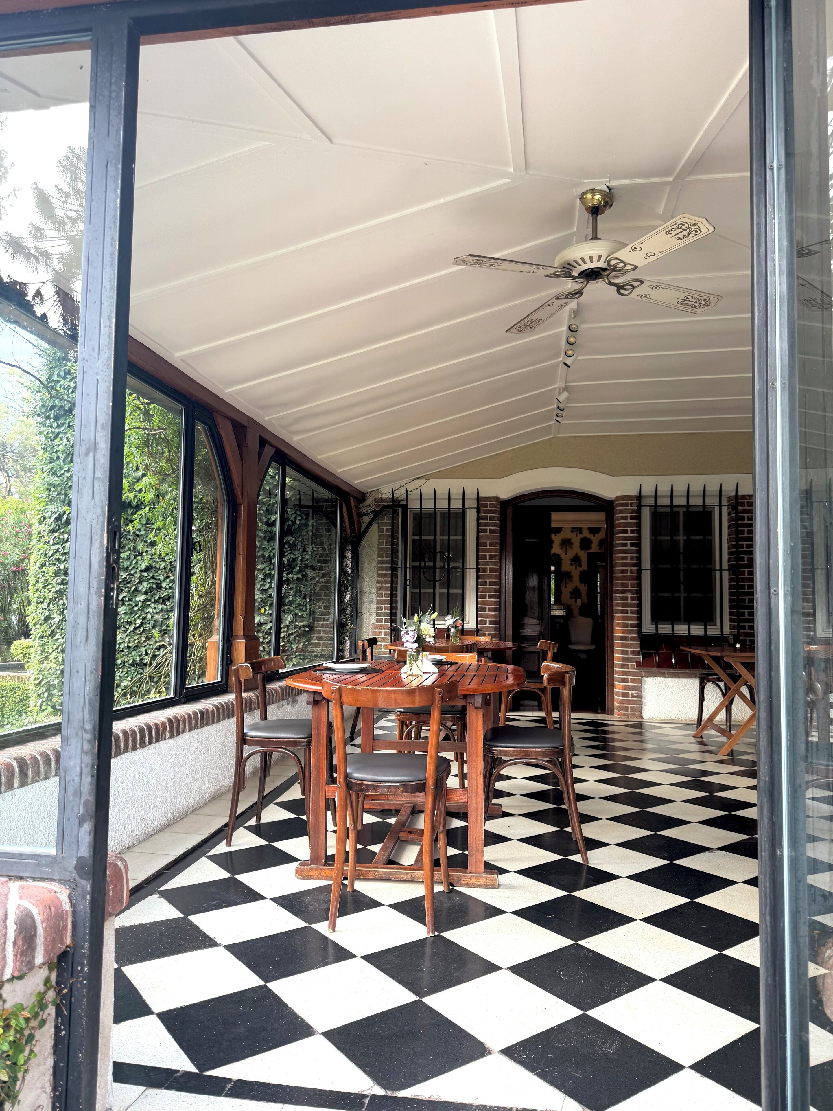 A covered outdoor seating area with wooden tables and chairs, a ceiling fan, and a checkered floor.