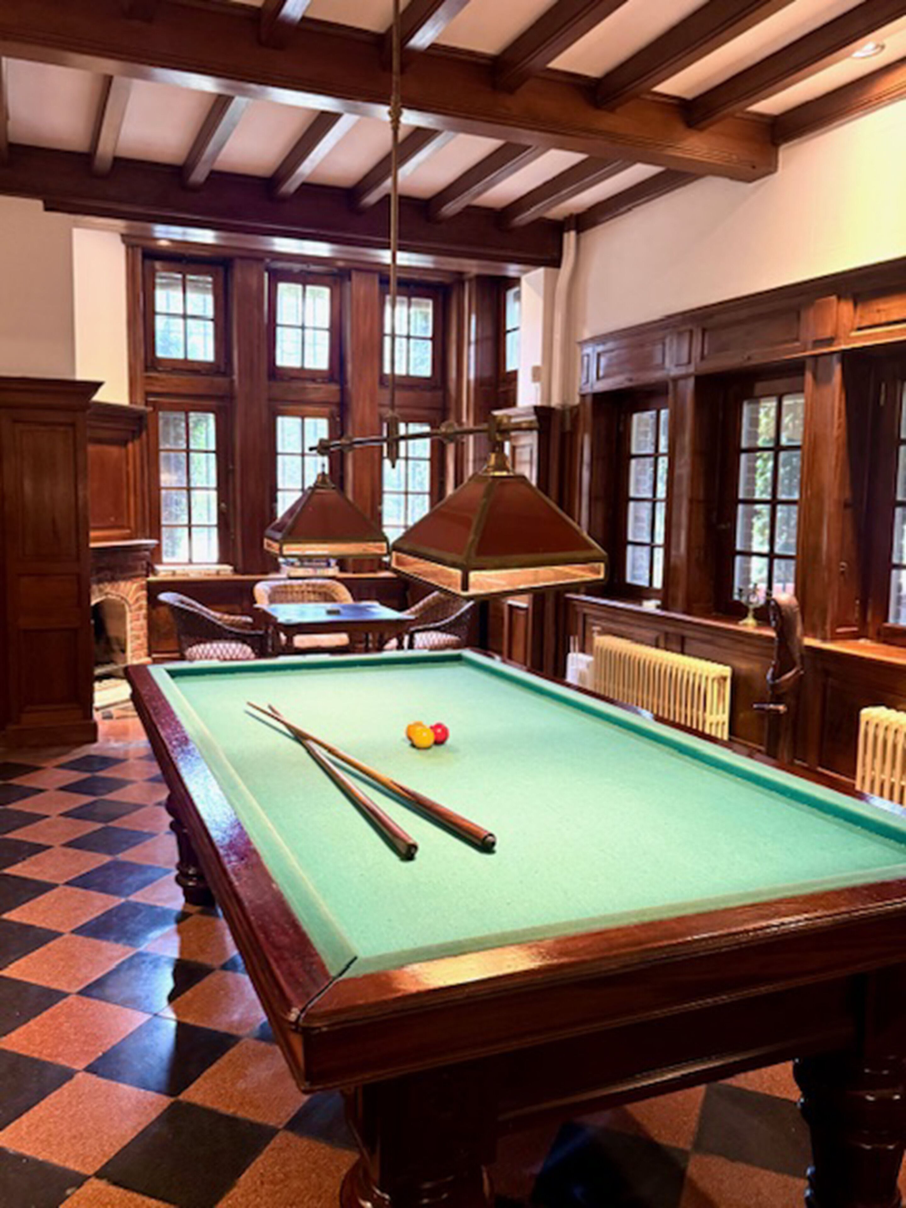A pool table with two cue sticks, a billiard ball, and a table lamp. There are wooden chairs and a small table with a lamp. The room has wooden beams on the ceiling and large windows with multiple panes.