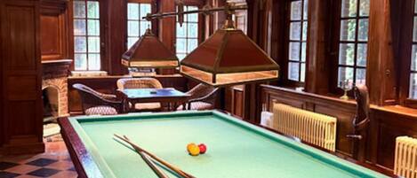A pool table with two cue sticks, a billiard ball, and a table lamp. There are wooden chairs and a small table with a lamp. The room has wooden beams on the ceiling and large windows with multiple panes.