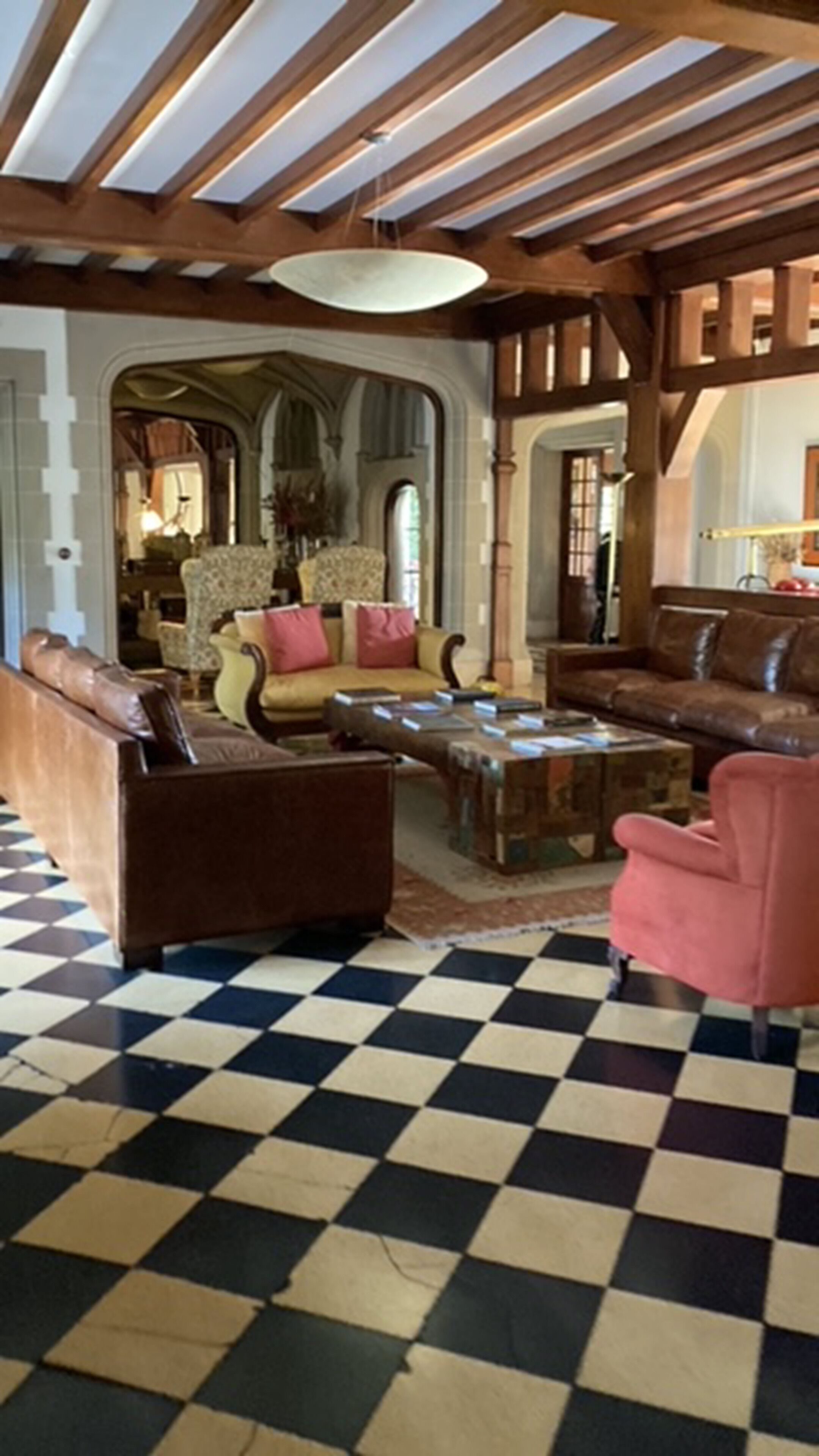 A spacious living room with a checkered floor, wooden ceiling beams, and a large chandelier.