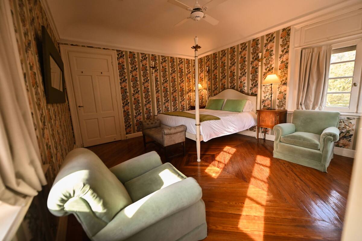 Quarto com cama de dossel, uma cadeira, mesa de cabeceira e janela com cortinas.