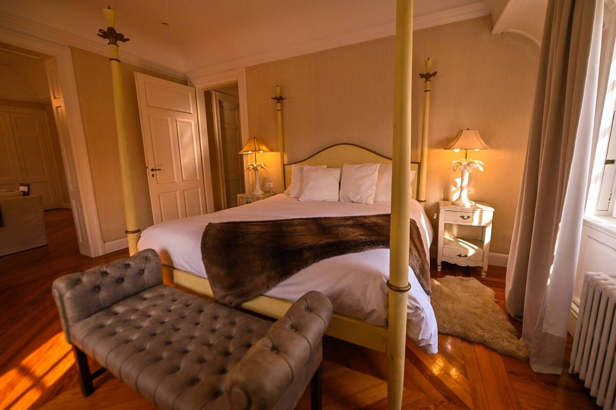 Uma cama de dossel com manta de pele, um banquinho estofado e duas mesas de cabeceira com abajures.