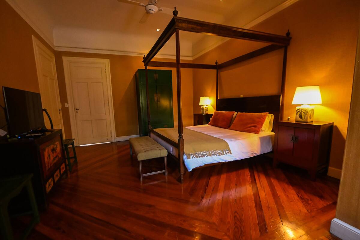 Uma cama de dossel com cabeceira de madeira, um banquinho, uma mesinha de cabeceira com abajur e uma televisão sobre um aparador.