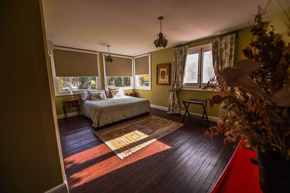 Um quarto com uma cama, uma mesa de cabeceira, uma janela com cortinas, piso de madeira e uma mesa lateral vermelha.
