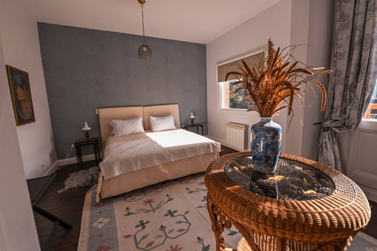 Quarto com cama, mesa de cabeceira, um vaso com plantas secas, janela com cortinas e um tapete estampado.