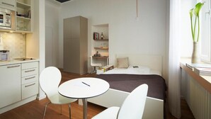 A modern hotel room with a bed, a desk, a chair, a small table, a kitchen area, and a window with a plant. - Aparthotel TU-TU (Essen)