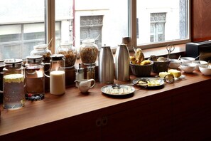 Un buffet de petit-déjeuner est installé sur un comptoir en bois près d’une fenêtre, avec une variété d’aliments et d’accompagnements.