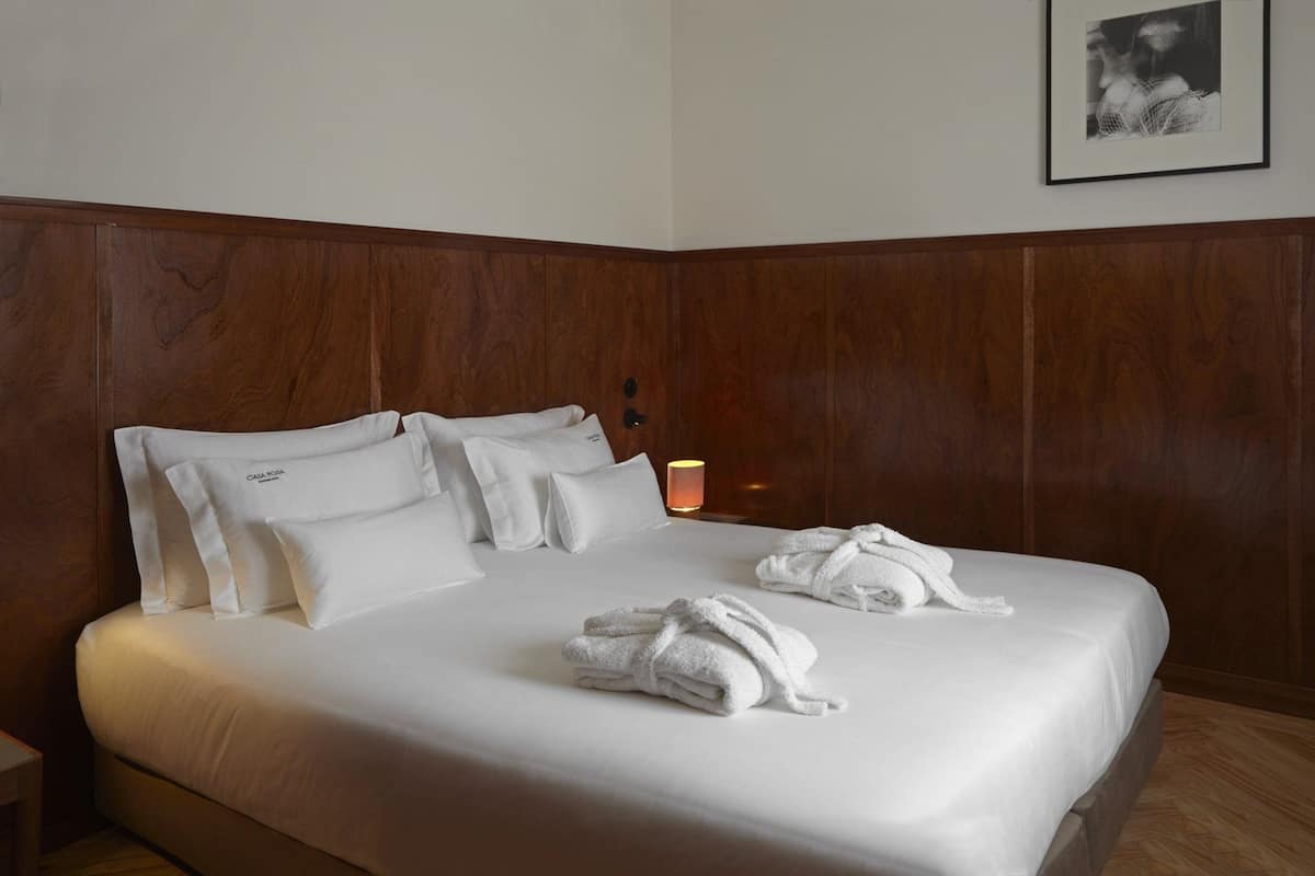 Una cama bien hecha con sábanas blancas y dos toallas enrolladas, una lámpara de noche y un cuadro en la pared.