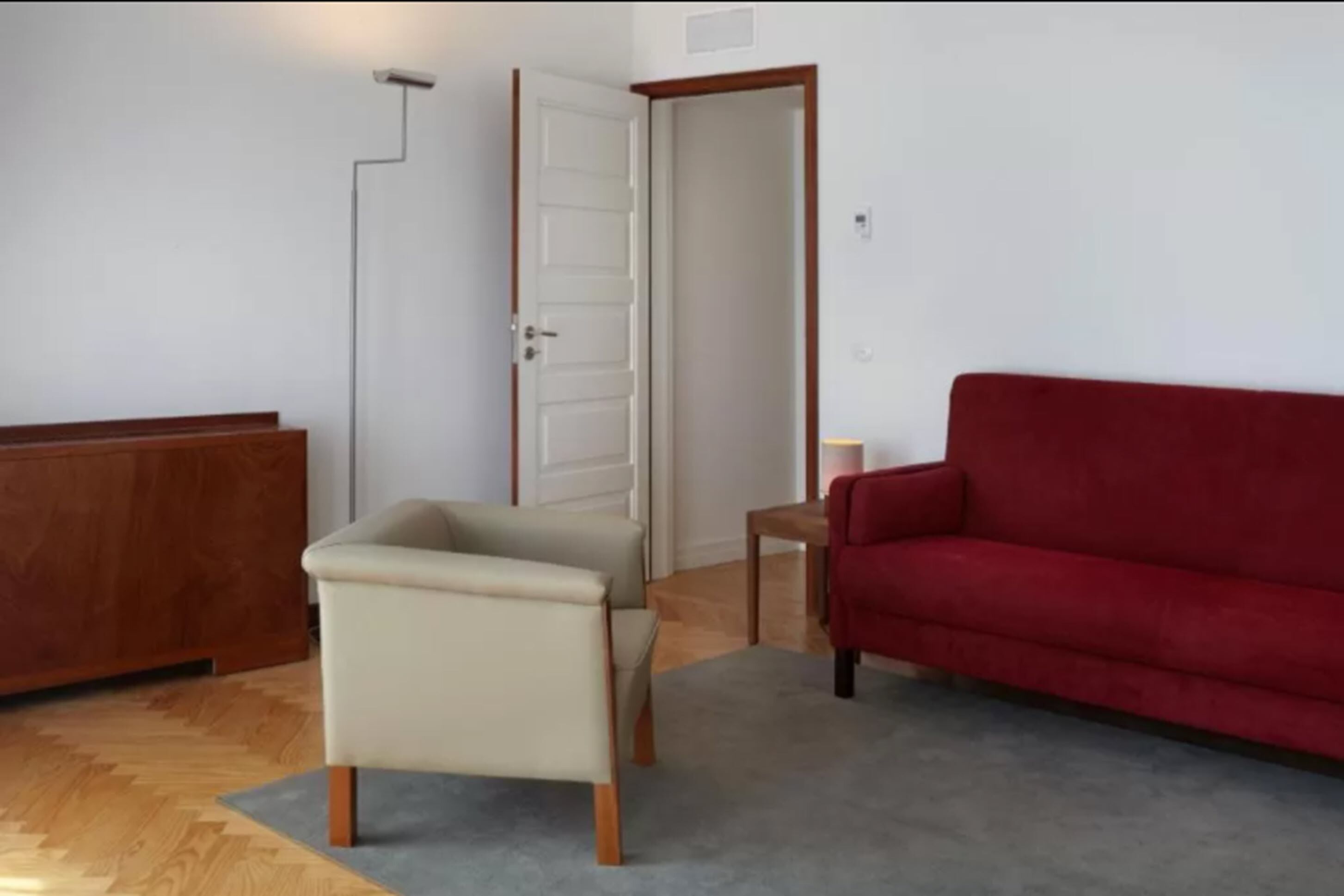 Une pièce avec un meuble en bois, un fauteuil beige, un canapé rouge et un tapis gris.