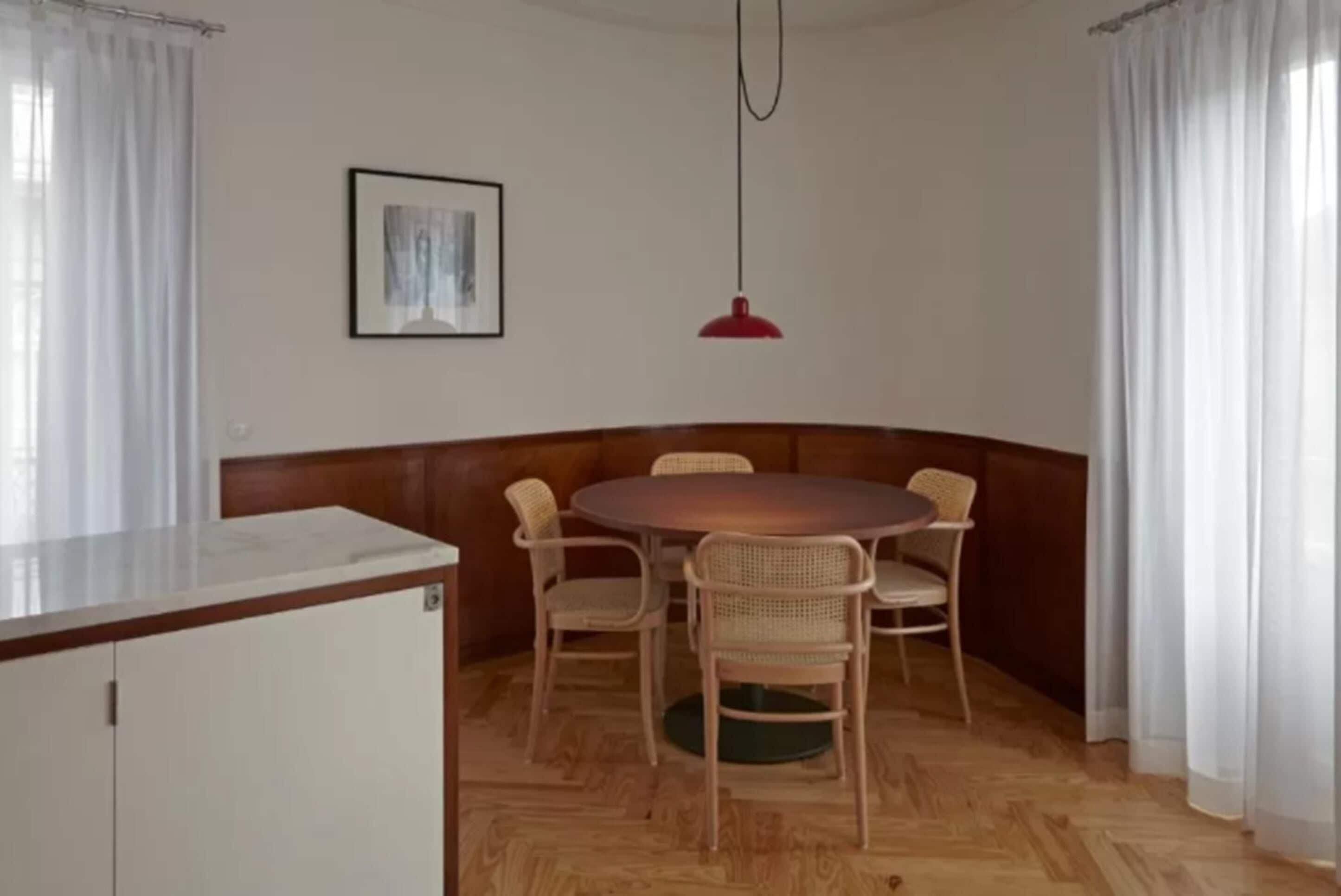 Un coin repas avec une table ronde et des chaises en osier, un luminaire suspendu rouge et un meuble blanc.