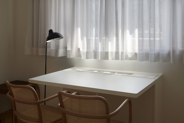 Une pièce avec un bureau blanc, deux fauteuils en osier, une lampe sur pied et des rideaux légers.