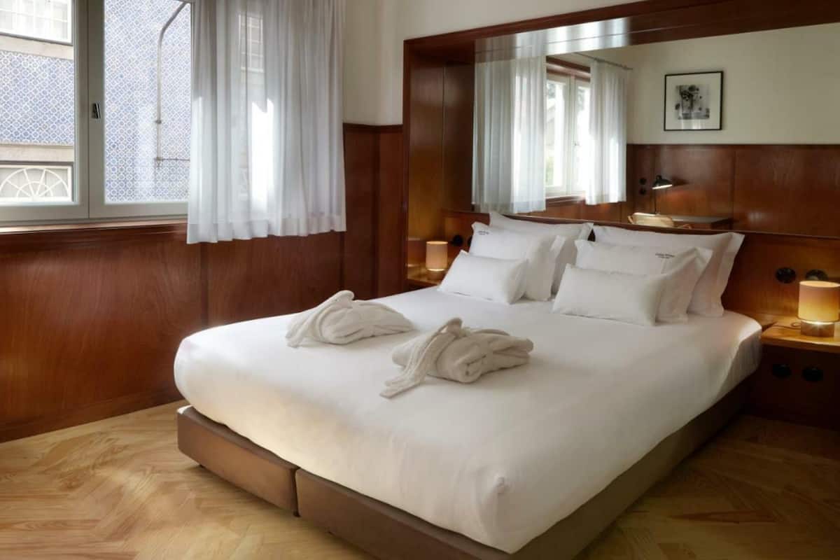 Una habitación de hotel con una cama grande, cabecera de madera, dos lámparas de noche y una ventana con cortinas.