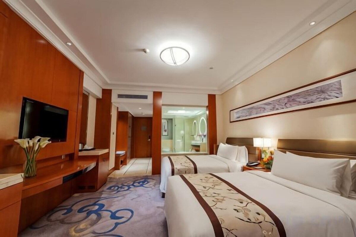 A hotel room with a television, a desk, two beds, a mirror, and a bathroom visible through an open door.