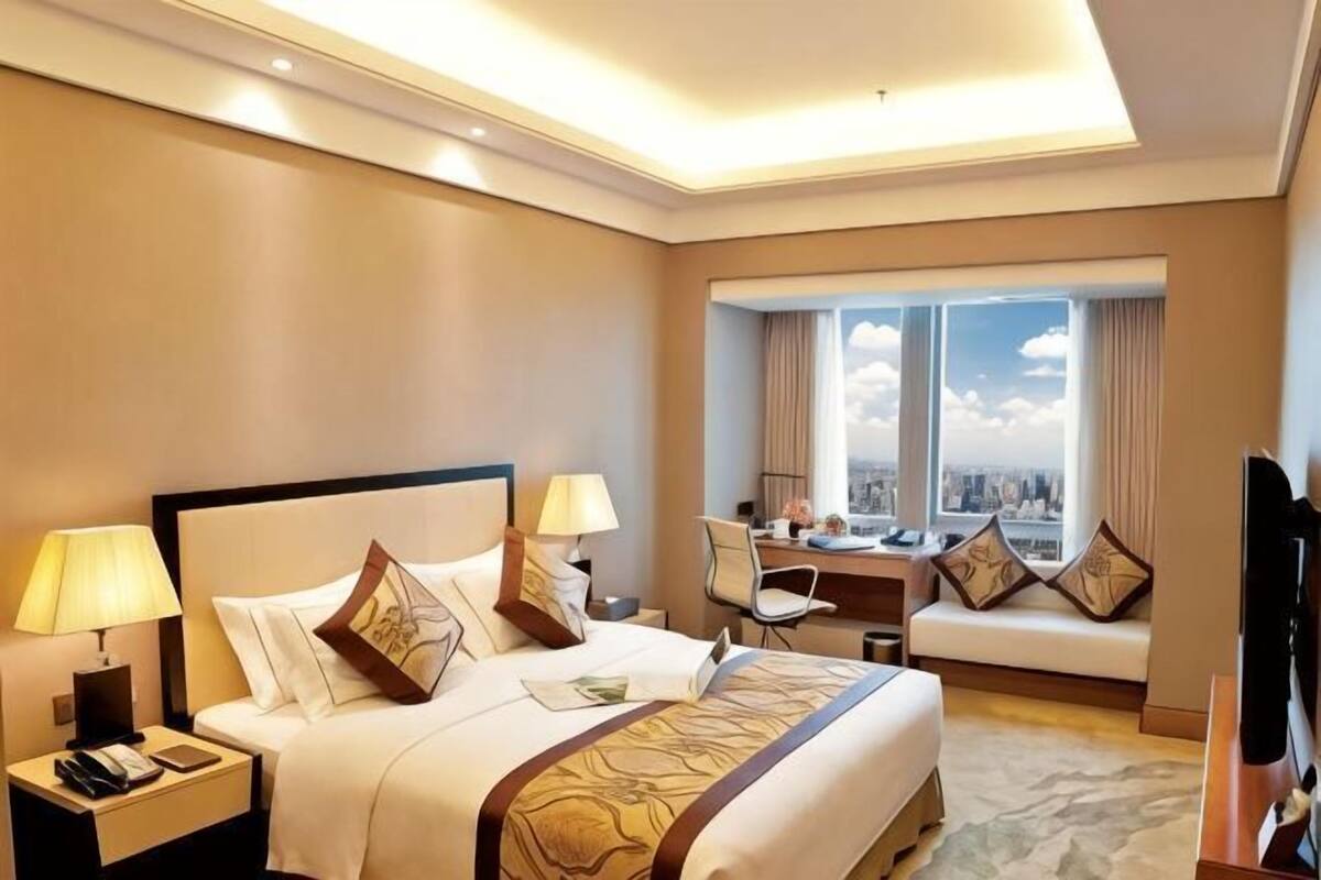 A hotel room with a bed, bedside lamps, a desk, a chair, a TV, and a large window with a city view.
