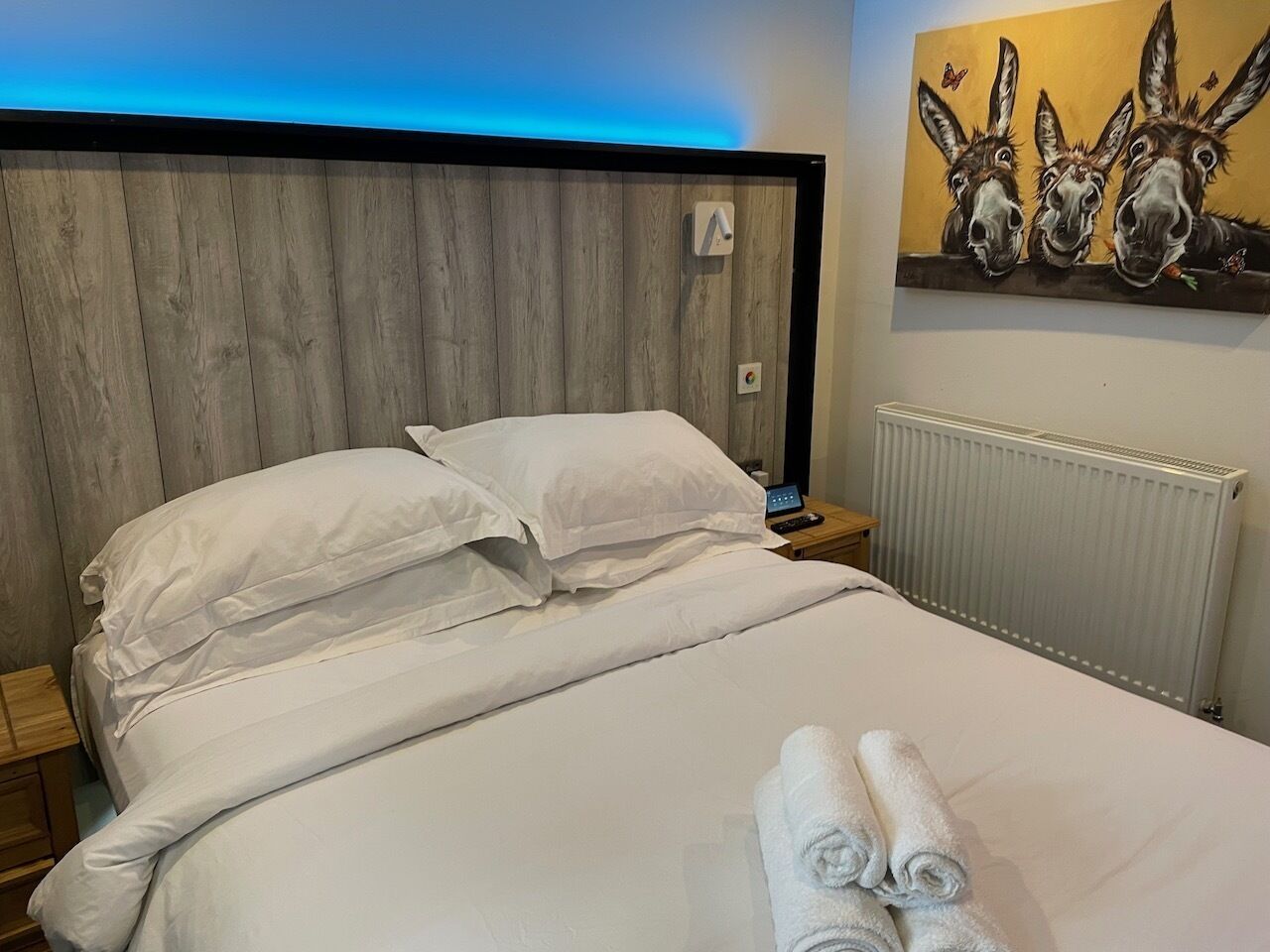 Una cama bien hecha con ropa de cama y toallas blancas, un cabecero de madera, un cuadro de burros en la pared y un termostato también en la pared.