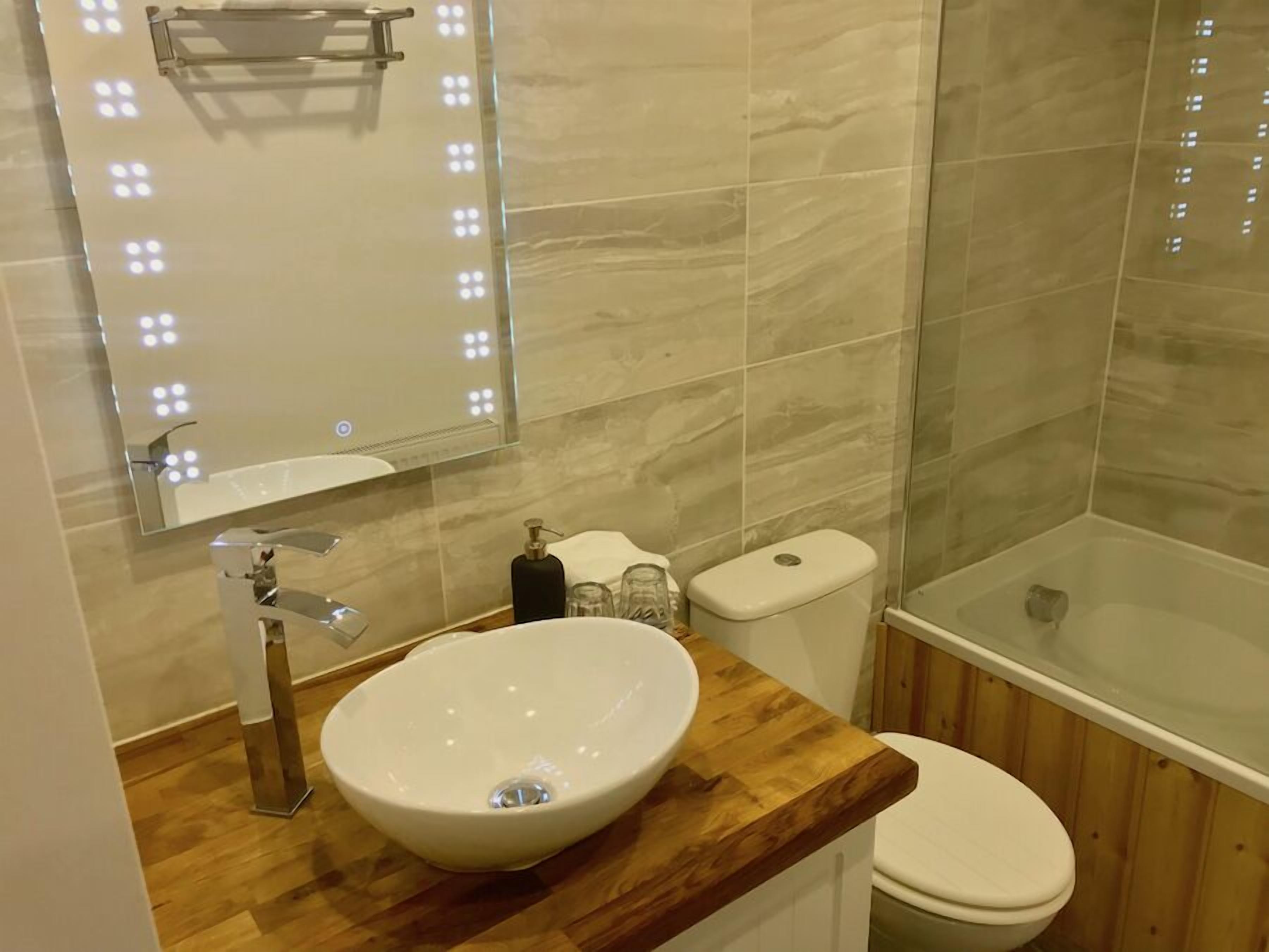 A bathroom with a wooden vanity, a white sink, a mirror with lights, a toilet, and a glass-enclosed shower.