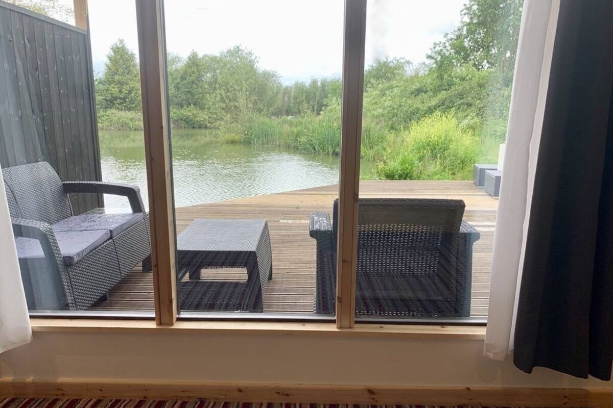 A room with a large window overlooking a wooden deck and a body of water. There are two wicker chairs and a small table on the deck.