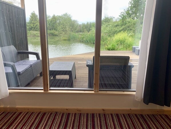 A room with a large window overlooking a wooden deck and a body of water. There are two wicker chairs and a small table on the deck. - The Waterfront Lodges (Bedworth)