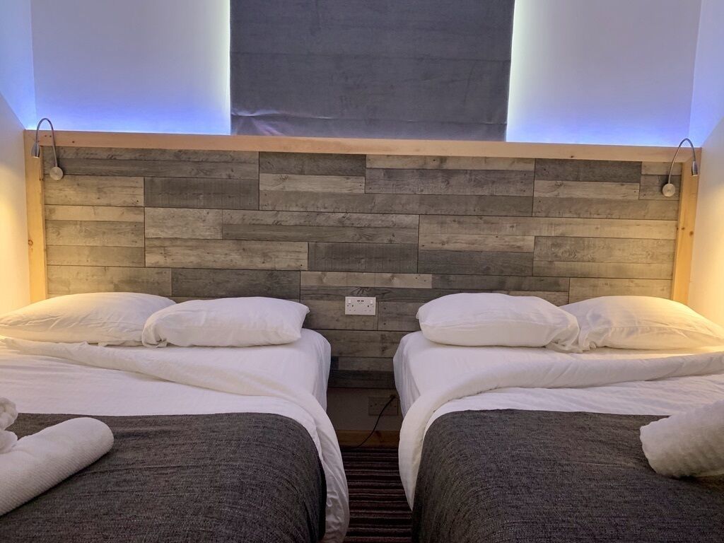 Habitación de hotel con dos camas, cada una con ropa blanca y una manta gris. El cabecero está hecho de listones de madera, y hay una ventana sobre las camas.