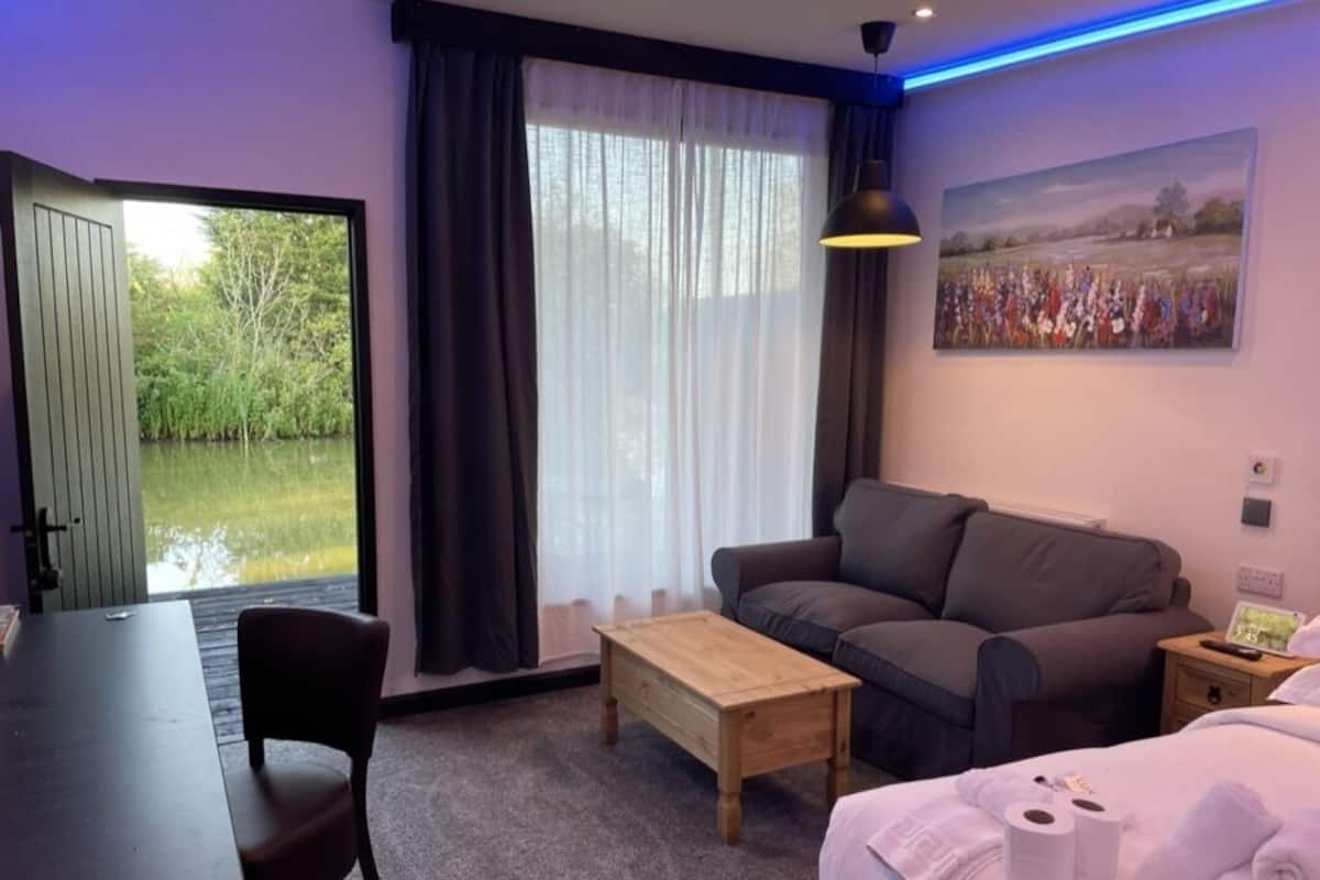 A modern hotel room with a large window, a sofa, a wooden table, and a bed with towels.