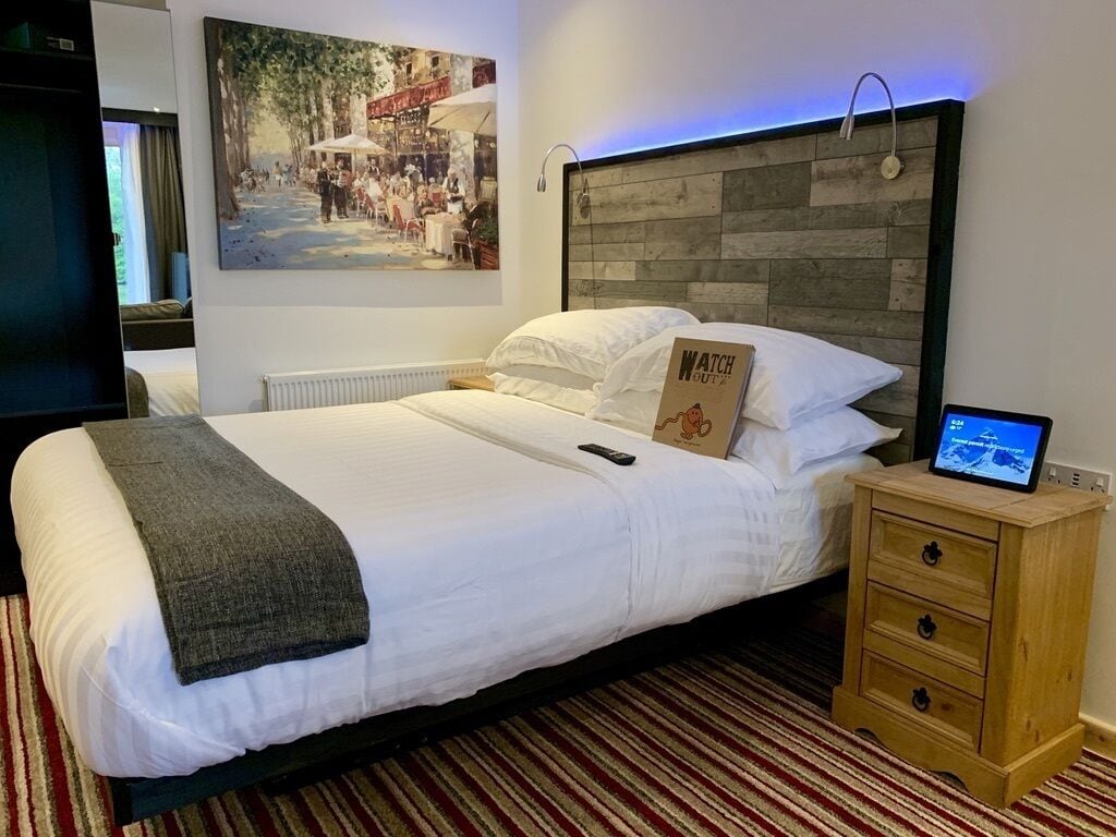 Una cama bien tendida con una manta gris, un cabecero de madera, una mesita de noche con una computadora portátil y un televisor montado en la pared.