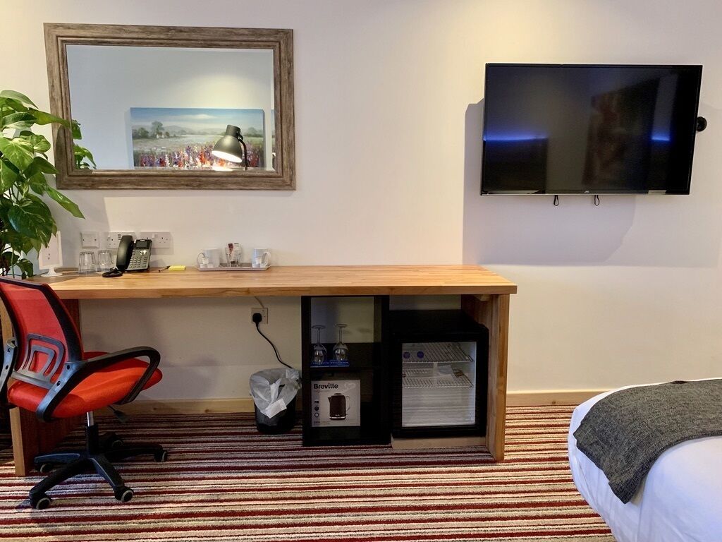A hotel room with a wooden desk, a red office chair, a television, a framed picture, and a bed with a blanket.