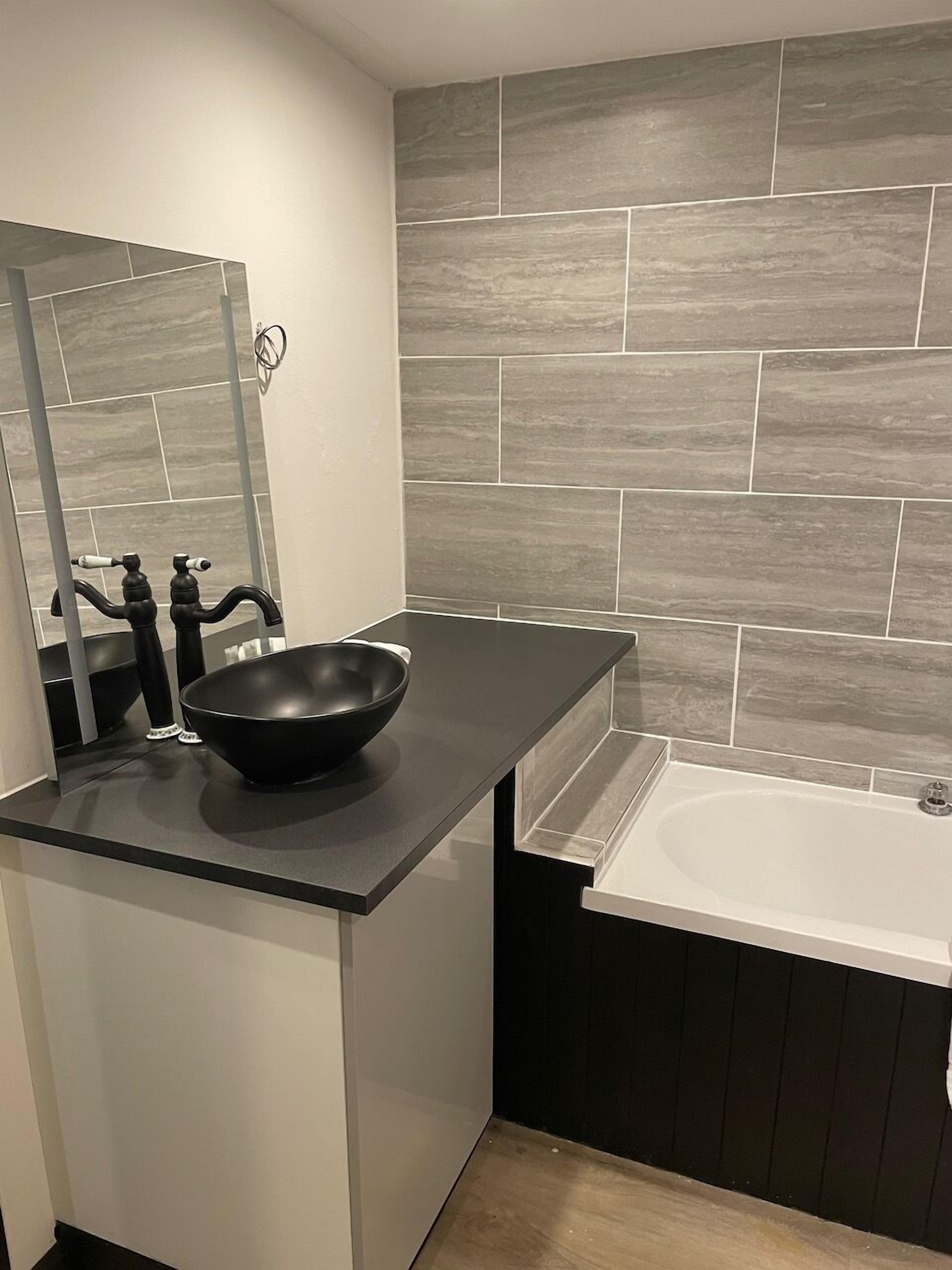 A modern bathroom with a black countertop, a black bowl sink, a white bathtub, and grey stone tiles on the wall.