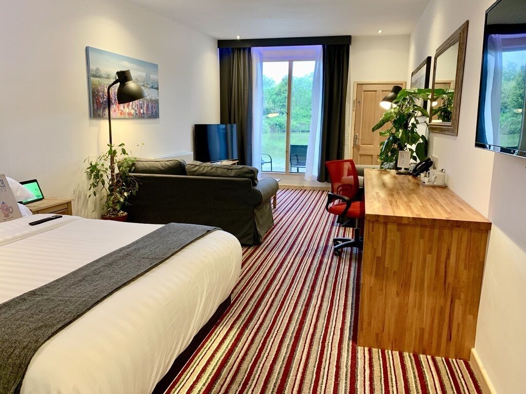 A hotel room with a bed, a desk, a sofa, a red chair, a plant, and a large window with a view of greenery.