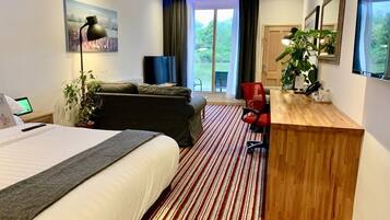 A hotel room with a bed, a desk, a sofa, a red chair, a plant, and a large window with a view of greenery.