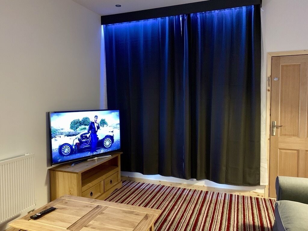 Uma sala de estar com televisão sobre um suporte de madeira, tapete listrado e uma janela grande com cortinas escuras.