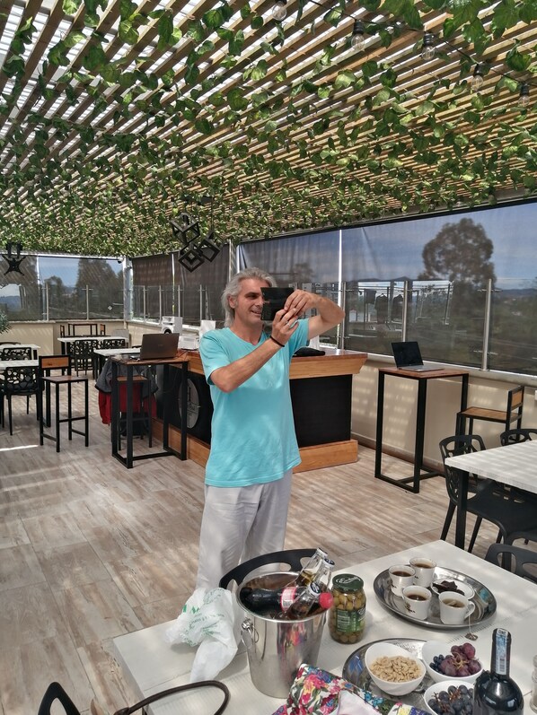 A person taking a selfie in a modern, well-lit outdoor dining area with a wooden floor, metal tables, and chairs.
