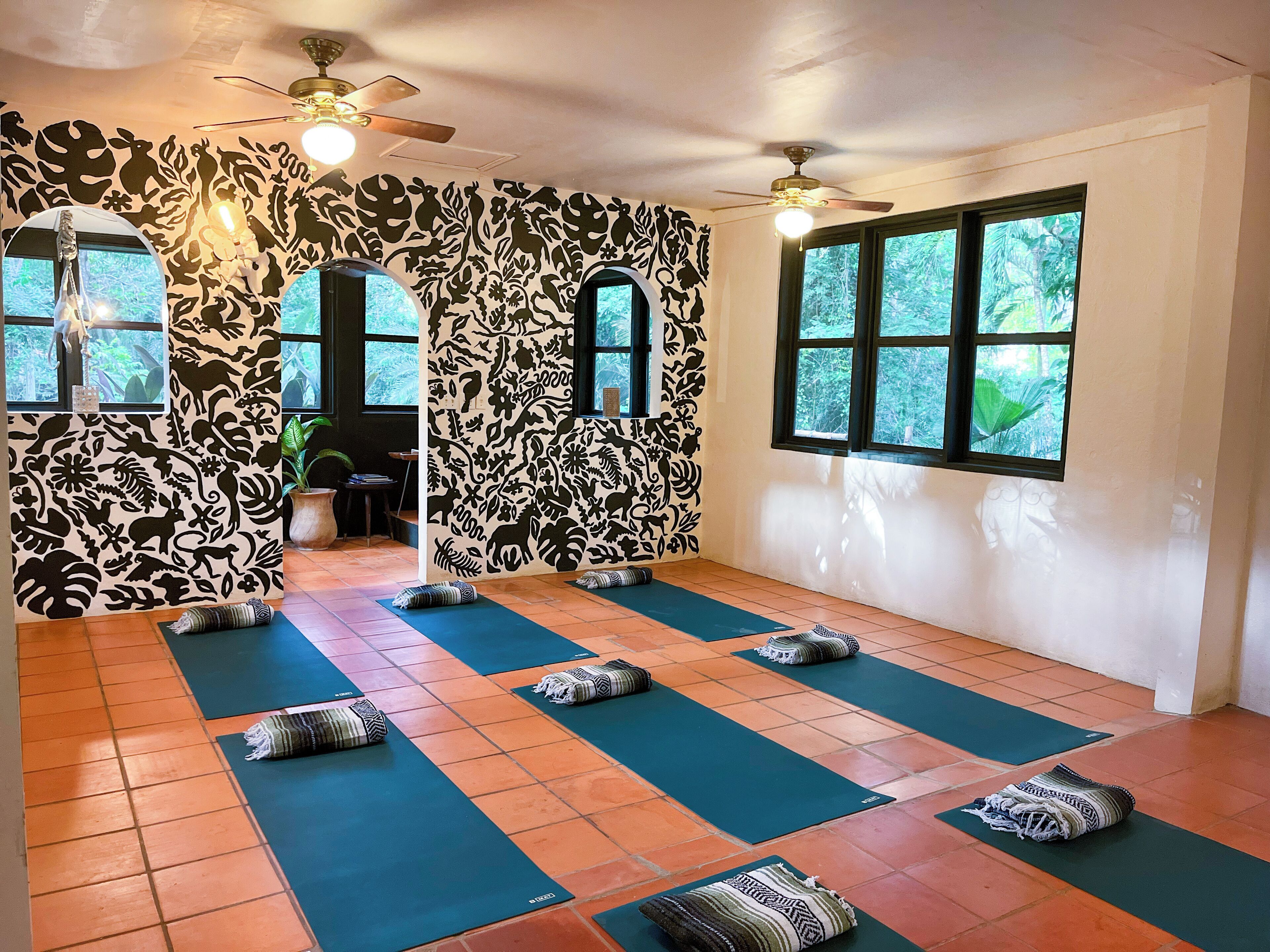 Un salón de yoga con paredes decoradas, ventanas amplias y numerosas colchonetas.