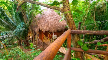Une hutte à toit de chaume, entourée d’une végétation tropicale luxuriante, reliée à une autre structure par un pont en bois.
