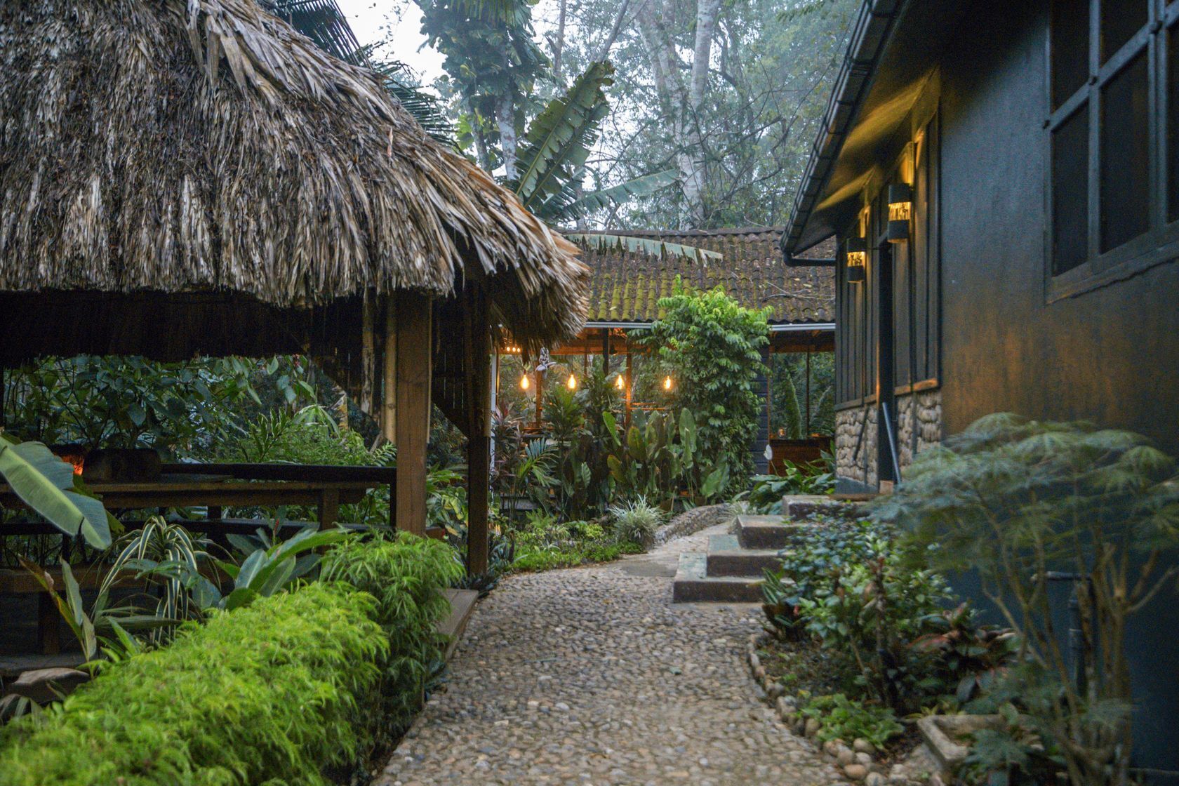 A rustic thatched-roof structure with a gravel pathway leading to a building with large windows.