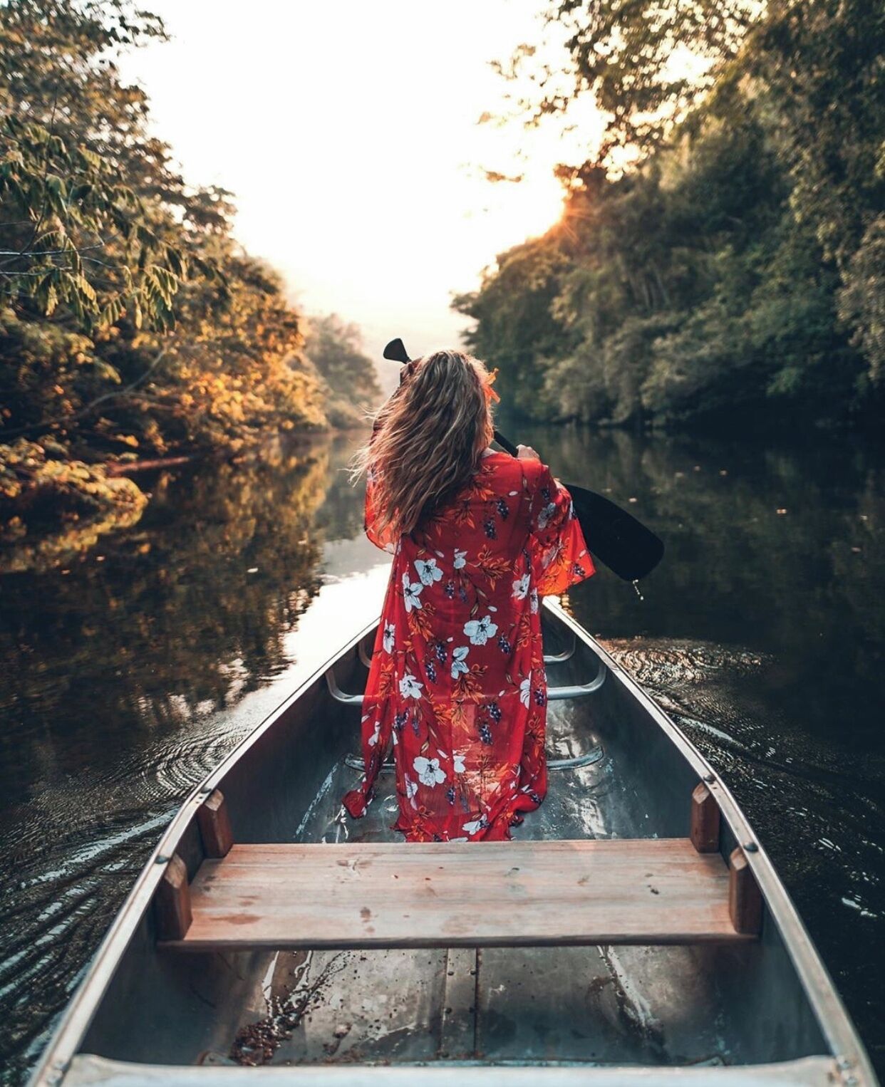 Eine Person in einem roten Blumenkleid paddelt mit einem Kanu einen ruhigen Fluss entlang, umgeben von Bäumen.