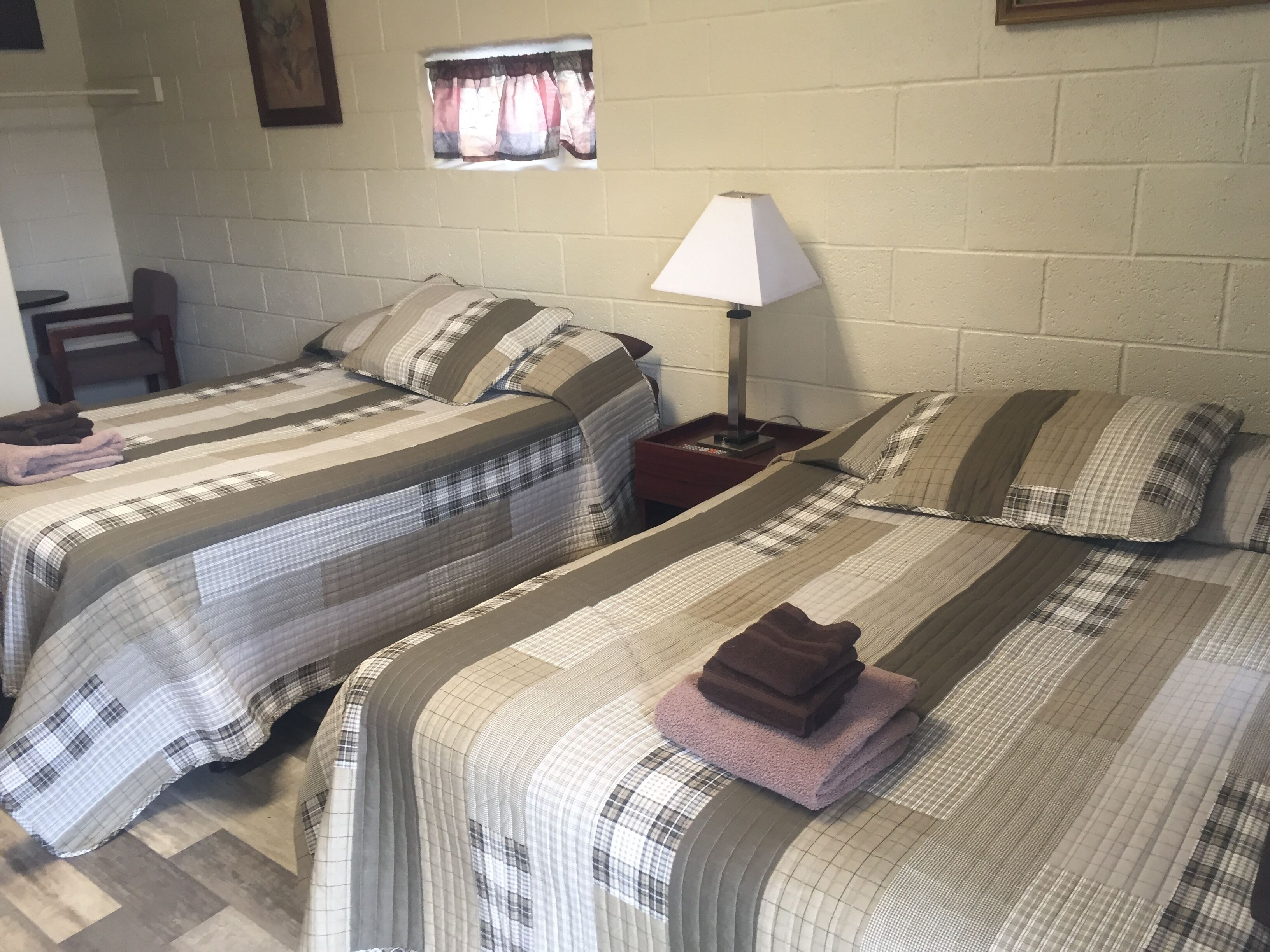 Ein Zimmer mit zwei Betten, jeweils mit gestreifter Bettwäsche, einem Nachttisch mit Lampe und einem kleinen Fenster mit Vorhängen.
