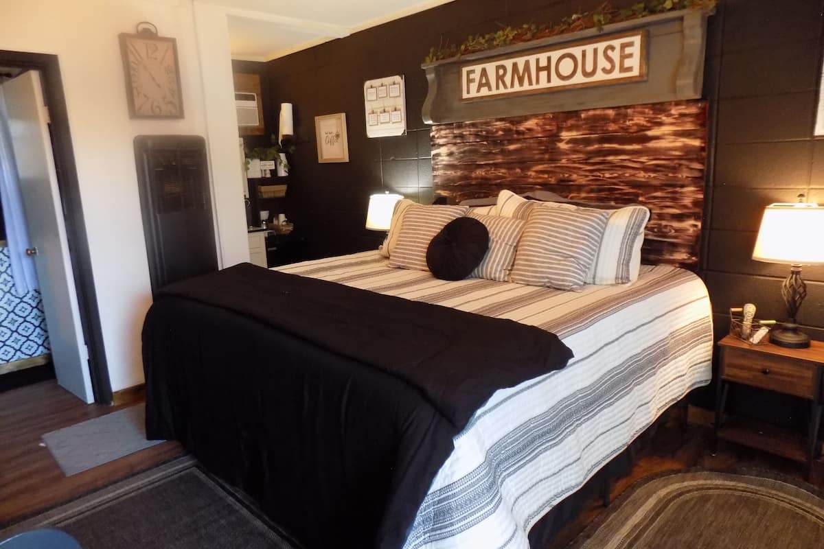 Quarto com cama, mesas de cabeceira, criado-mudo, relógio na parede, placa com a inscrição 'FARMHOUSE' e uma porta.