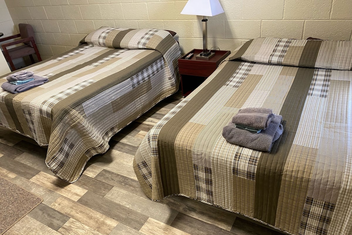 Quarto com duas camas, cada uma com colcha xadrez e toalha dobrada no lado direito. Um criado-mudo com abajur fica entre as camas.